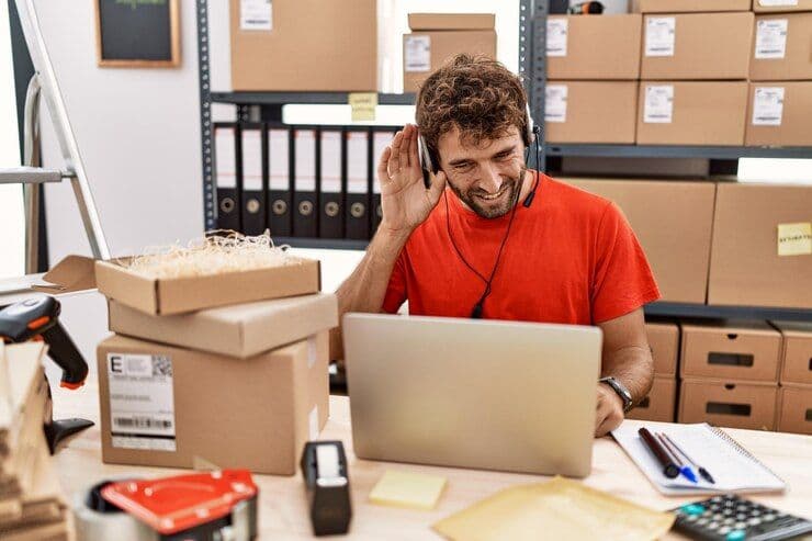 A young man in a red shirt, wearing a headset, is working on a laptop in a busy shipping office surrounded by boxes and packing supplies.