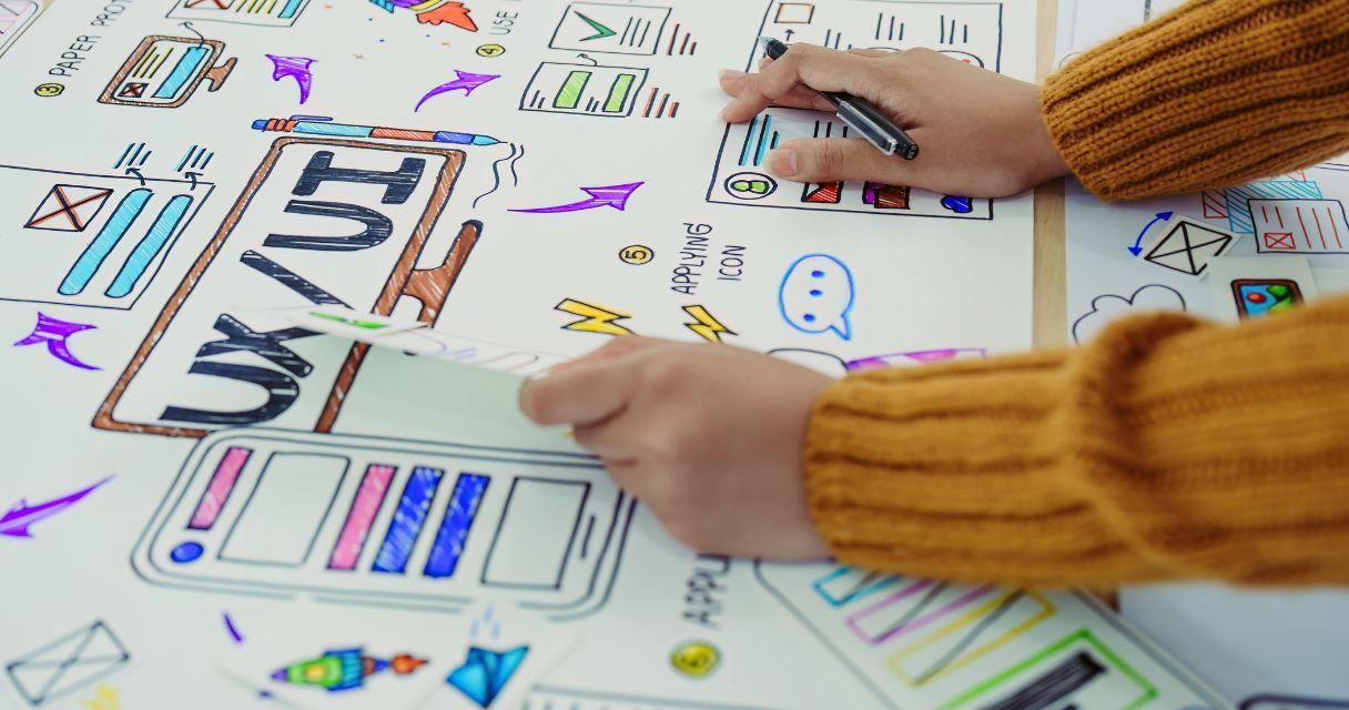 The image shows a person working on a colorful UX/UI design layout with sketches and notes on a large paper.