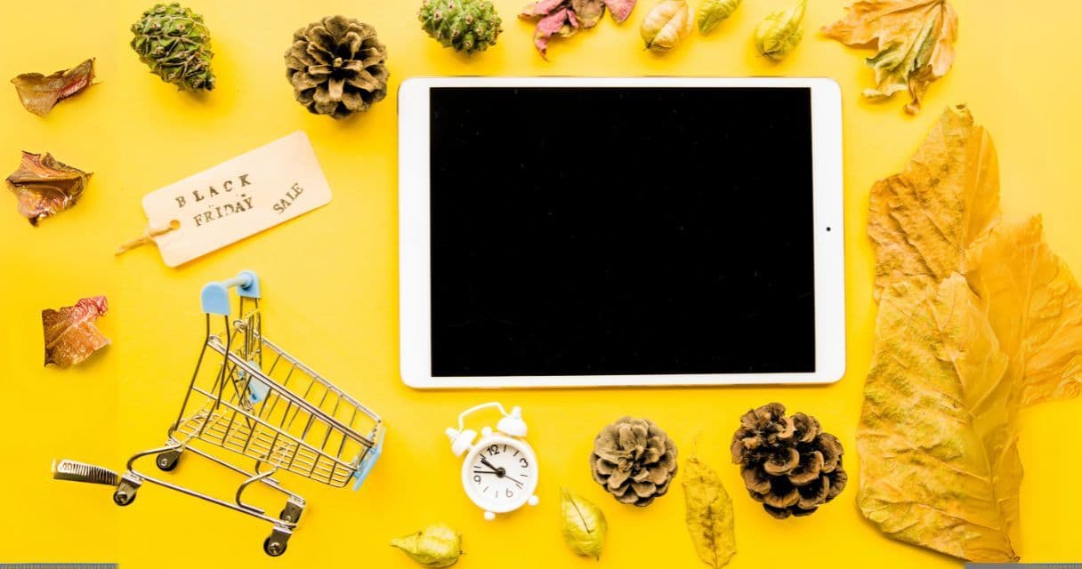 This image depicts a bright yellow background with a tablet, a small shopping cart, a "Black Friday Sale" tag, a white alarm clock, and scattered autumn leaves and pine cones, symbolizing holiday shopping and seasonal promotions.